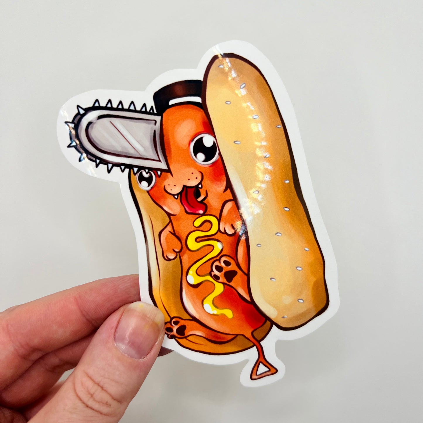 A hand holds a die-cut sticker featuring a cartoon hot dog character with a chainsaw head, mustard, and a bun, against a plain light background.
