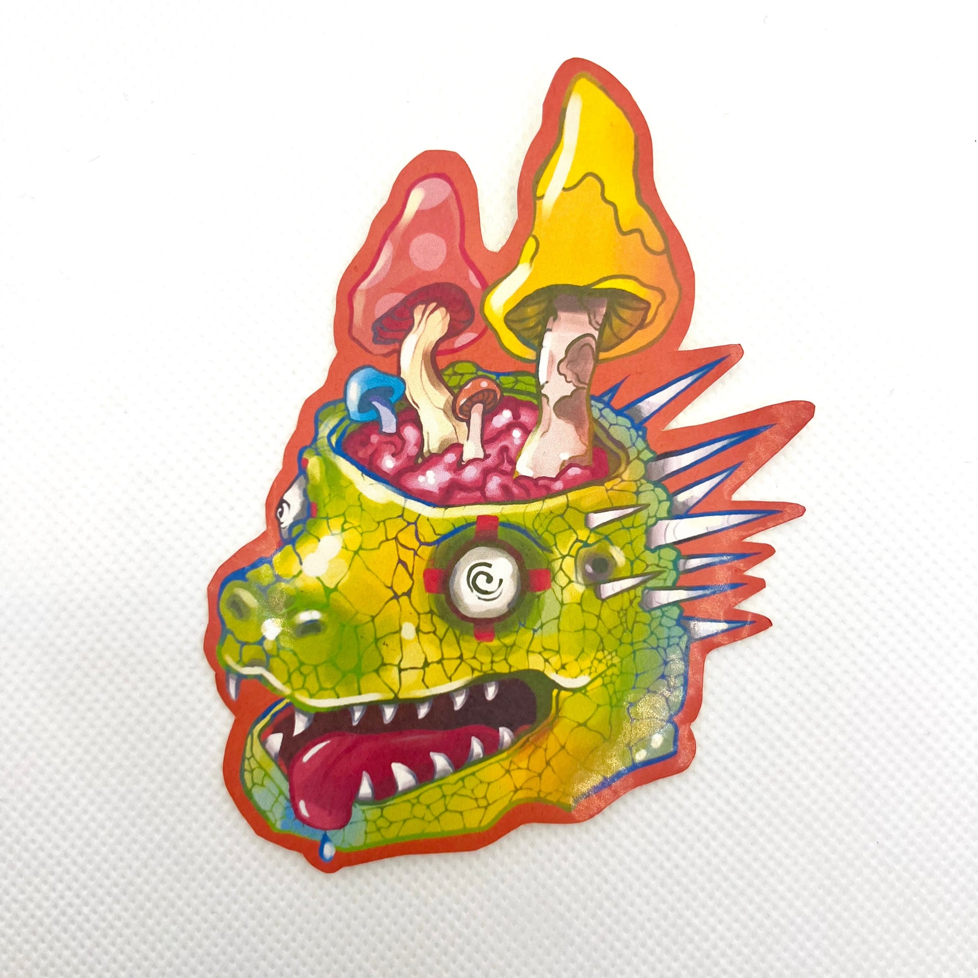 A colorful die-cut sticker or patch featuring a green reptile-like head with mushrooms growing out of it, shown on a plain white textured background.