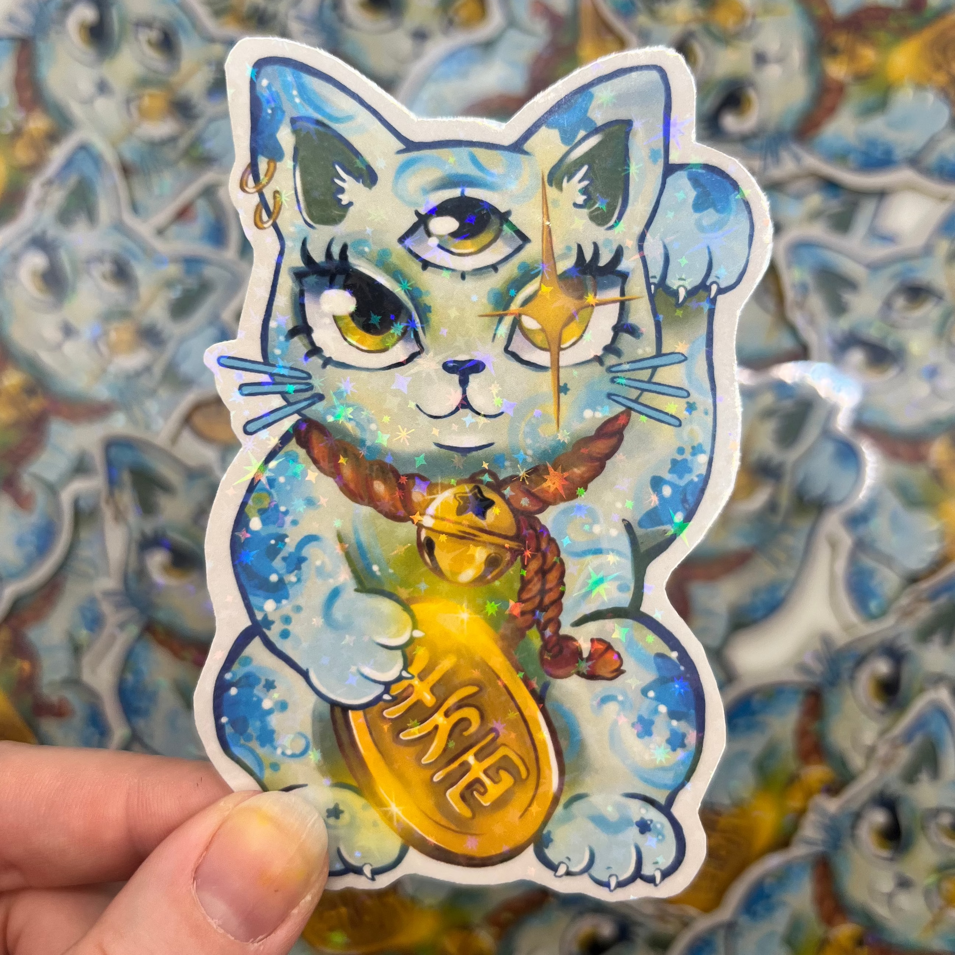 A hand holds a die-cut sticker of a blue and white cat holding a gold ingot, with a blurred background of multiple identical stickers.