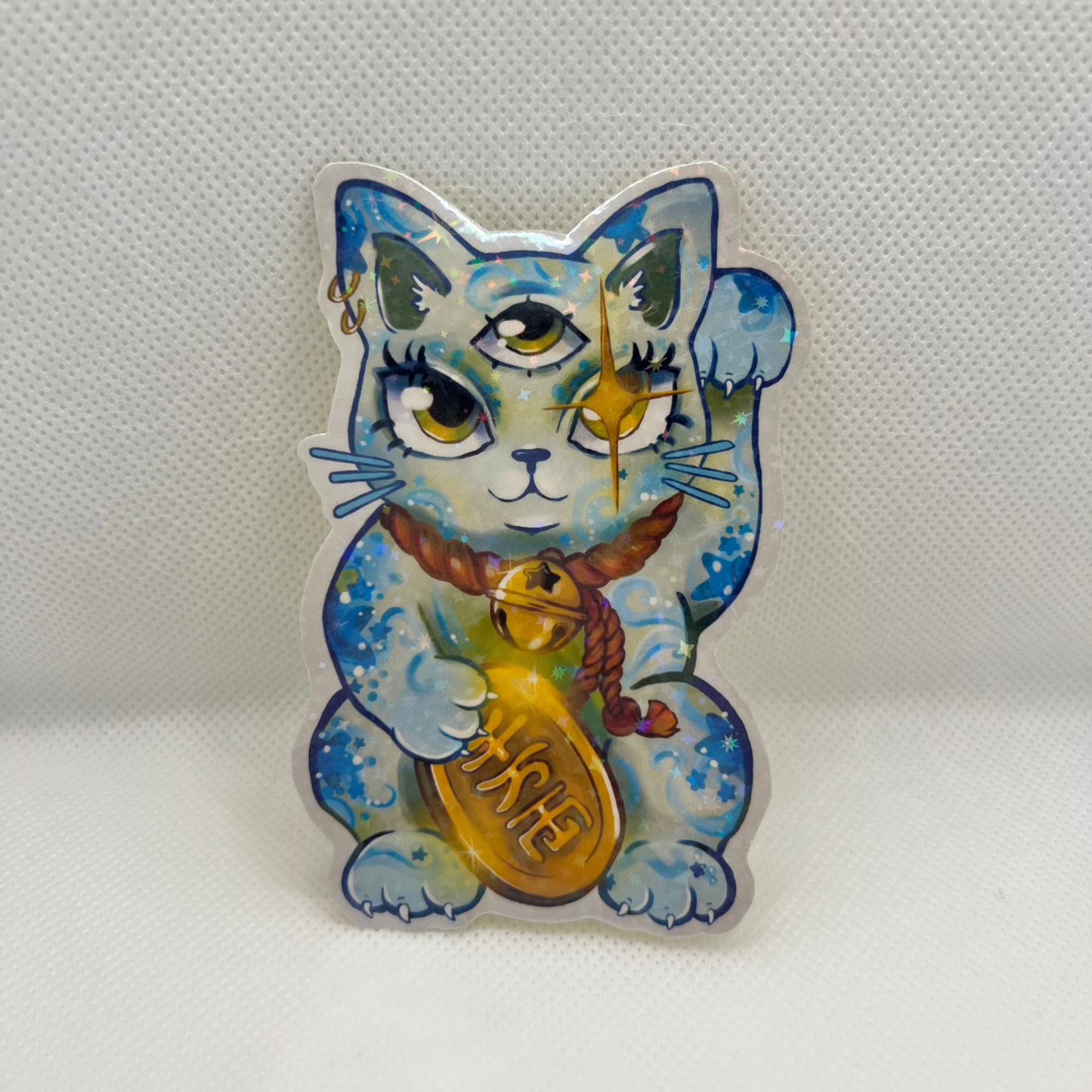 A die-cut acrylic pin featuring a blue and white cat illustration with gold accents, holding a gold bell, set against a textured white background.