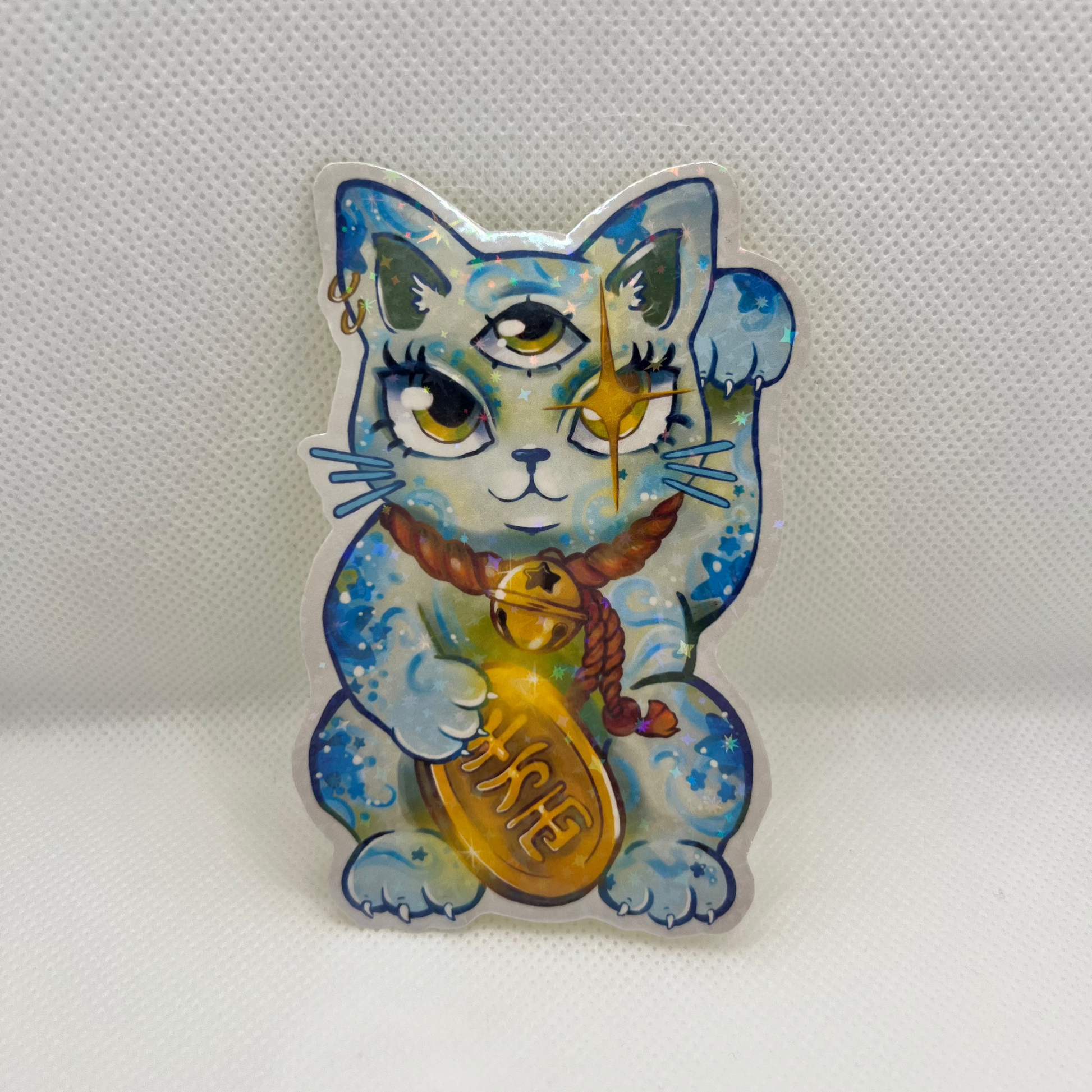 A die-cut acrylic pin featuring a blue and white cat illustration with gold accents, holding a gold bell, set against a textured white background.